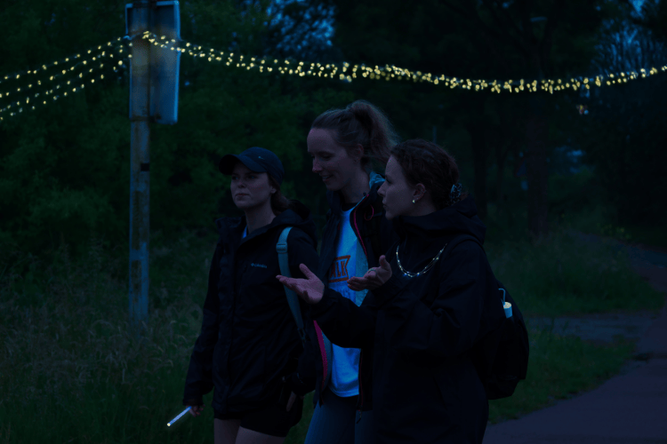 Drie wandelende vrouwen in de nacht met lichtslinger op de achtergrond.